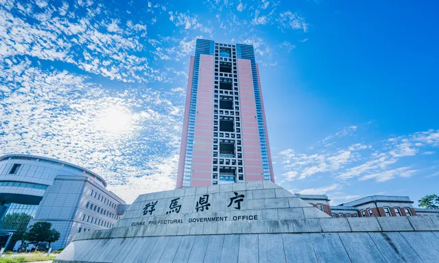 Tall government office building for Gunma Prefecture with a stone podium displaying the sign 'Gunma Prefectural Government Office' against a bright blue sky.